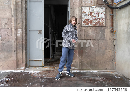 Young boy standing outdoors by an open metal door of a stone building. Winter street portrait Young boy standing outdoors by an open metal door of a stone building. Winter street portrait 137543558