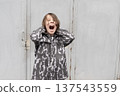 Young boy screaming with hands on head. Frustrated child shouting outdoors against grey metal door 137543559