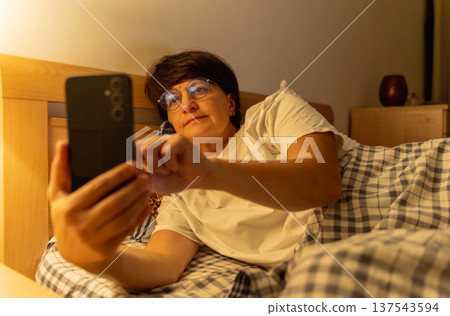 Mature sleepy woman lying in bed browsing social media on smartphone at night, insomnia, internet addiction Mature sleepy woman lying in bed browsing social media on smartphone at night, insomnia, internet addiction 137543594
