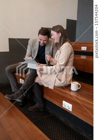 Young woman and man in business attire talking and looking at paperwork together on wooden benches. Attire, conversation, paperwork, workspace, business. 137544204