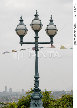 A commanding, ornate Victorian lamppost stands prominently, featuring a dark green post and three distinct, orb-like lanterns. Each lantern, encased in a frame and topped with a matching green finial. A commanding, ornate Victorian lamppost stands prominently, featuring a dark green post and three distinct, orb-like lanterns. Each lantern, encased in a frame and topped with a matching green finial. 137544242