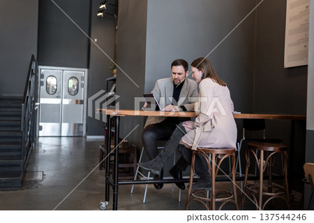 Man and woman working at high cafe table, using laptops together in open modern workspace. High table, laptops, modern workspace, teamwork, business. Man and woman working at high cafe table, using laptops together in open modern workspace. High table, laptops, modern workspace, teamwork, business. 137544246