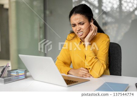 Young businesswoman suffering from severe earache and an ear infection, touching her painful ear while struggling to concentrate on her work at a modern office desk with a laptop 137544774