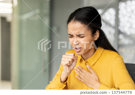 Young woman suffering from a cough and sore throat, showing symptoms of flu or cold in an office environment, experiencing discomfort and pain from sickness 137544779