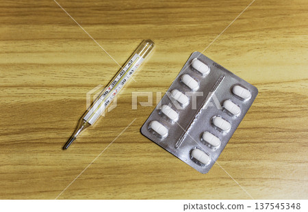 Medical thermometer and a blister pack of white pills on a wooden table. Medical thermometer and a blister pack of white pills on a wooden table. 137545348