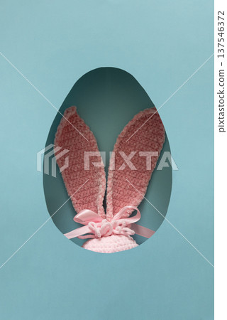 Pink Easter plush bunny ears peeking through oval egg shaped cutout in pastel blue background Pink Easter plush bunny ears peeking through oval egg shaped cutout in pastel blue background 137546372
