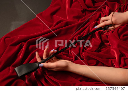 Female hands with manicured nails holding a black leather riding crop on luxurious red velvet fabric. 137546407