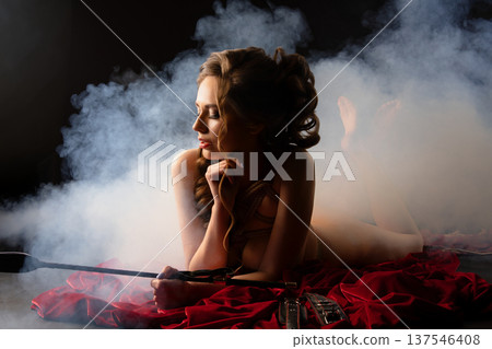 Elegant woman in shibari rope tie holding a riding crop, lying on red velvet in dramatic smoke. 137546408