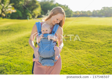 Mother using ergonomic baby carrier during a walk with her infant outdoors. Comfortable babywearing, active motherhood lifestyle, bonding, modern parenting, hands free care and everyday family routine 137546507