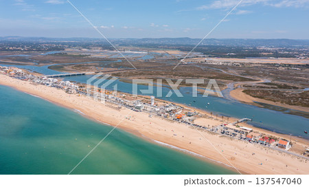Aerial view shows Praia de Faro with people on the beach and Ria Formosa nearby. The coastline stretches as boats are visible in the water on a clear day Aerial view shows Praia de Faro with people on the beach and Ria Formosa nearby. The coastline stretches as boats are visible in the water on a clear day 137547040
