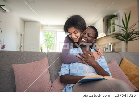 Young Woman Embracing Her Mother Reading a Book on the Sofa at Home Young Woman Embracing Her Mother Reading a Book on the Sofa at Home 137547329