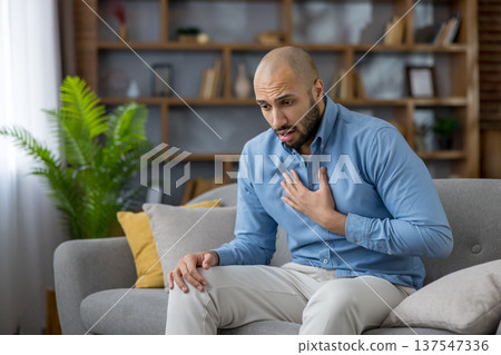 Man experiencing acute chest pain while sitting on a sofa in his living room, struggling with a potential heart attack or respiratory issues, needing immediate medical attention Man experiencing acute chest pain while sitting on a sofa in his living room, struggling with a potential heart attack or respiratory issues, needing immediate medical attention 137547336