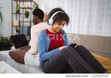 Two Young Women Sitting Back to Back on Sofa, One Working on Laptop, Other Listening to Music 137547337