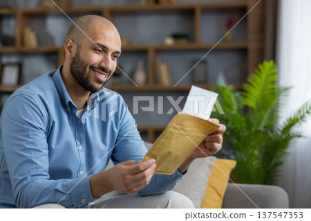 Smiling man on a sofa reads good news from a brown envelope, pulling out a letter in a modern living room, relaxed and pleased as he opens personal mail at home 137547353