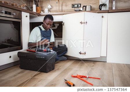 Plumber writing on clipboard while inspecting pipes under kitchen sink Plumber writing on clipboard while inspecting pipes under kitchen sink 137547364