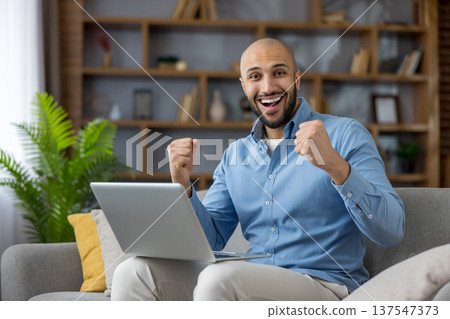 Smiling man raising fists in triumph, sitting on a sofa with a laptop, celebrating achievement and good news while working remotely in a comfortable home office setting Smiling man raising fists in triumph, sitting on a sofa with a laptop, celebrating achievement and good news while working remotely in a comfortable home office setting 137547373