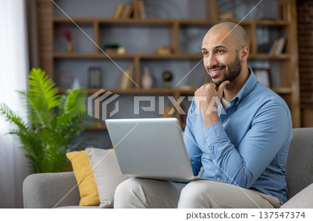 Smiling young man relaxing on a sofa while telecommuting with his laptop, focused and confident remote worker studying or freelancing from a comfortable modern home office Smiling young man relaxing on a sofa while telecommuting with his laptop, focused and confident remote worker studying or freelancing from a comfortable modern home office 137547374
