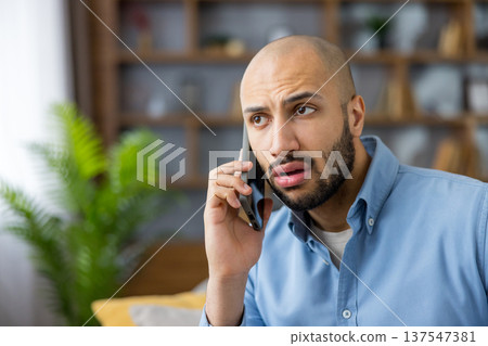 Bald man with beard listening to a surprising or troubling phone conversation, showing a reaction of doubt and concern while holding a mobile device indoors Bald man with beard listening to a surprising or troubling phone conversation, showing a reaction of doubt and concern while holding a mobile device indoors 137547381
