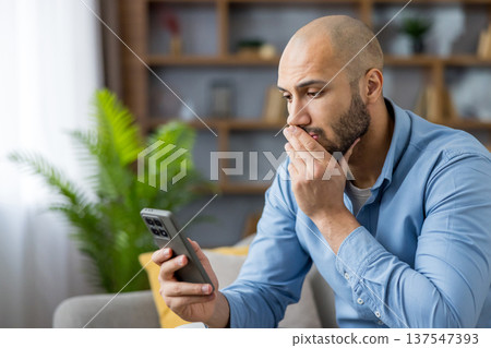 Young bald man looking worried at his mobile phone, covering his mouth with a hand, sitting on a sofa in a modern apartment, processing shocking or upsetting information Young bald man looking worried at his mobile phone, covering his mouth with a hand, sitting on a sofa in a modern apartment, processing shocking or upsetting information 137547393