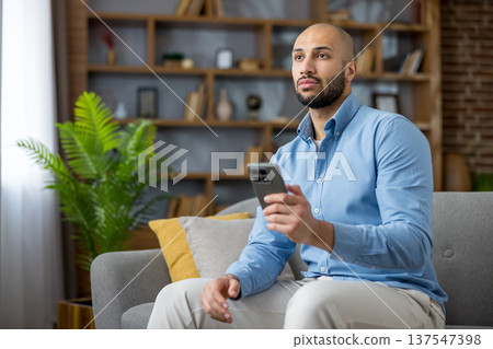 Thoughtful man sitting on a sofa, holding a mobile phone, and looking away, pondering new ideas or remote work solutions in a comfortable living room setting 137547398