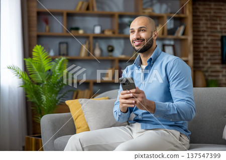 Smiling young man sitting comfortably on sofa in modern living room, holding phone and looking away with thoughtful expression, enjoying digital communication and connectivity 137547399