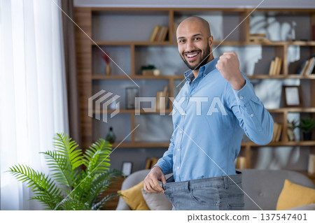 Man proudly celebrating his successful weight loss journey, clenching a fist in triumph and showing off the loose fit of his old, oversized jeans at home 137547401