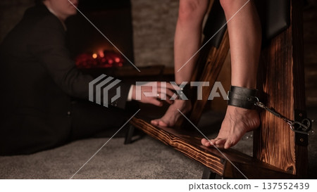 A man chains a woman's legs to a BDSM cross. A man chains a woman's legs to a BDSM cross. 137552439