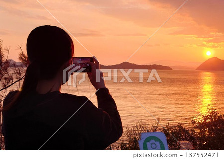 Back view of a woman taking a photo of the sunset with her smartphone Back view of a woman taking a photo of the sunset with her smartphone 137552471