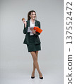Happy young business woman in a dark green suit standing full length on a white background, holding an orange folder with papers and a coffee cup, looking away. Curly hair, brown-haired 137552472