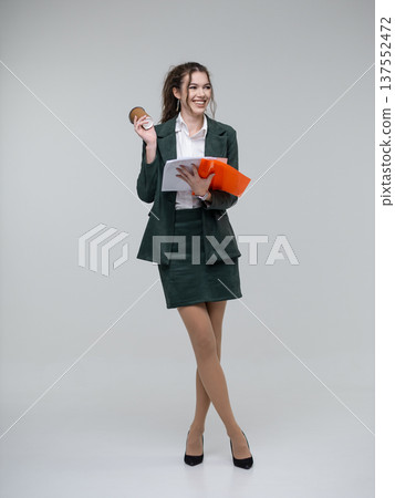Happy young business woman in a dark green suit standing full length on a white background, holding an orange folder with papers and a coffee cup, looking away. Curly hair, brown-haired 137552472