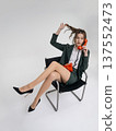 An elegant white woman in a green business suit talks on a red telephone while sitting on a chair against the backdrop of an isolated space. Office. Curly hair, brown-haired 137552473