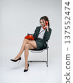 An elegant white woman in a green business suit talks on a red telephone while sitting on a chair against the backdrop of an isolated space. Office. Curly hair, brown-haired 137552474