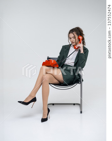 An elegant white woman in a green business suit talks on a red telephone while sitting on a chair against the backdrop of an isolated space. Office. Curly hair, brown-haired An elegant white woman in a green business suit talks on a red telephone while sitting on a chair against the backdrop of an isolated space. Office. Curly hair, brown-haired 137552474