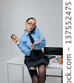 Young white woman in office attire is talking on the phone holding a cup of coffee and a folder with documents She is sitting on the edge of table in business attire Blue shirt glasses Isolated space 137552475