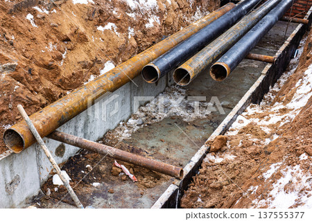 Excavated trench with large rusty and black water pipes ready for installation near new building construction site Excavated trench with large rusty and black water pipes ready for installation near new building construction site 137555377
