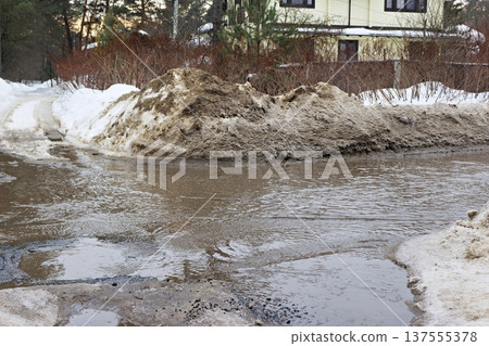 Melting dirty snow piles flank a muddy stream flowing across a road surface near a suburban dwelling Melting dirty snow piles flank a muddy stream flowing across a road surface near a suburban dwelling 137555378