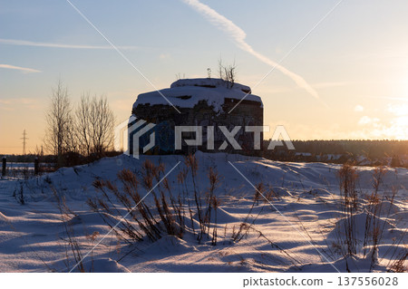 Graffiti-covered old brick bunker sits atop a snowy hill at sunset, winter landscape with dry grass in foreground 137556028