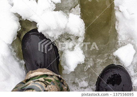 Heavy rubber boots breaking through fragile, melting river ice. The perilous surface reveals dangerous cold water, a clear warning of late winter hazards and unsafe conditions Heavy rubber boots breaking through fragile, melting river ice. The perilous surface reveals dangerous cold water, a clear warning of late winter hazards and unsafe conditions 137556095