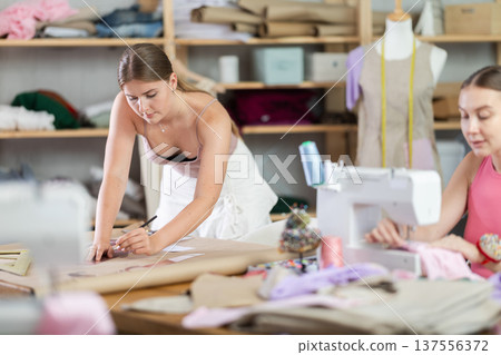 Female dressmakers working in the workshop against the background of racks with fabrics 137556372