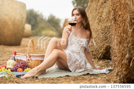 Woman in white dress drinking red wine on picnic near hay bales 137556595