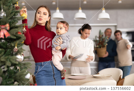 Young woman with a baby in her arms decorates a Christmas tree with toys while the rest of the family prepares festive dinner 137556602