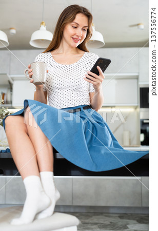 Cheerful woman in trendy dress typing message on mobile phone screen in kitchen 137556774