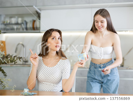 Young woman giving money to her sister in kitchen Young woman giving money to her sister in kitchen 137556869
