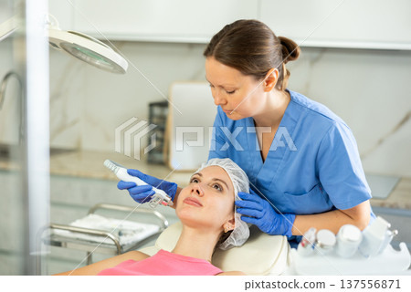Beautiful female doctor making facial gas-liquid oxygen water epidermal peeling using professional equipment use macromolecule atomizing pen for young girl at spa salon. Skin care concept. Beauty Beautiful female doctor making facial gas-liquid oxygen water epidermal peeling using professional equipment use macromolecule atomizing pen for young girl at spa salon. Skin care concept. Beauty 137556871