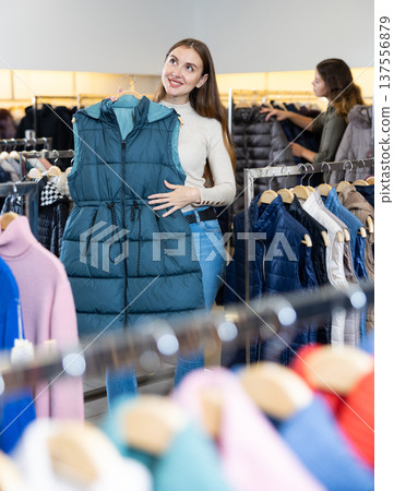 Dreamy young woman holding turquoise autumn vest while choosing clothes among the racks in a mall 137556879