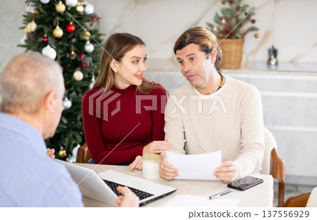 Male realtor discussing home choice with couple in real estate agency office, offering various options for residence against the background of Christmas tree 137556929