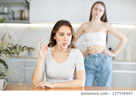 Angry mother sitting at the kitchen table, daughter standing behind Angry mother sitting at the kitchen table, daughter standing behind 137556947