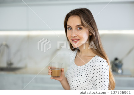 Girl is sitting on kitchen table and holding clear glass cup with latte coffee in hands 137556961