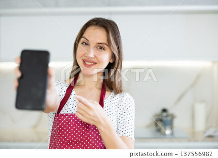 At home in kitchen smiling woman in apron shows blank smartphone screen close up. Dark touch screen workspace 137556982