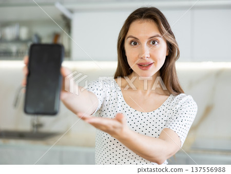 In kitchen, smiling girl in casual clothes shows blank smartphone screen close-up 137556988